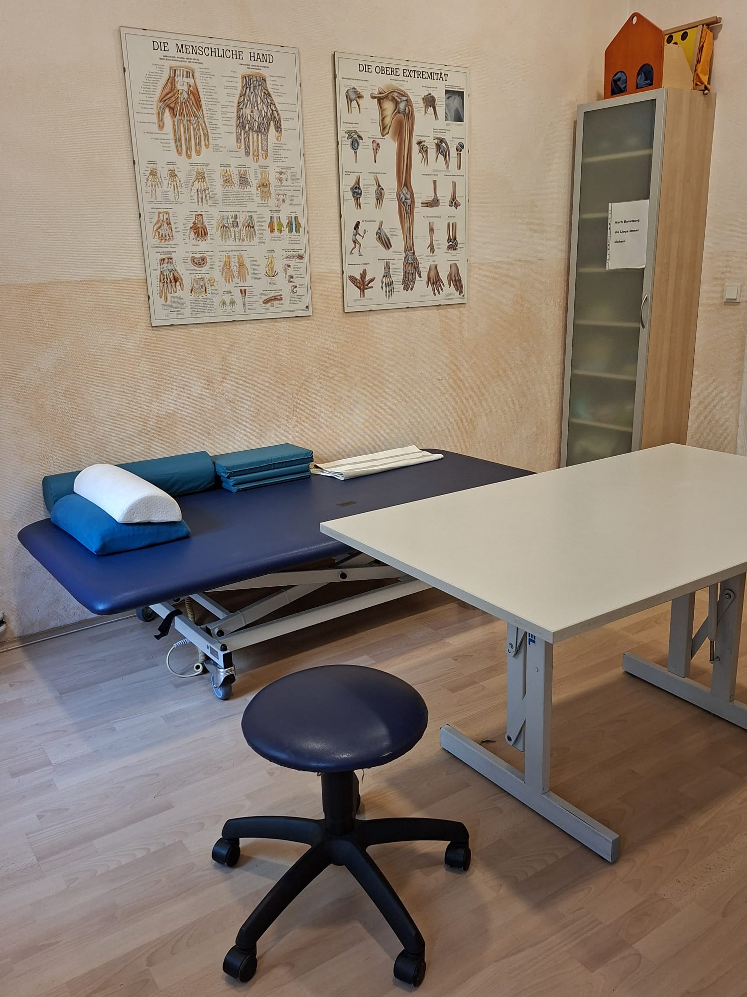 Behandlungsraum mit Therapieliege, Arbeitstisch und Anatomie-Postern